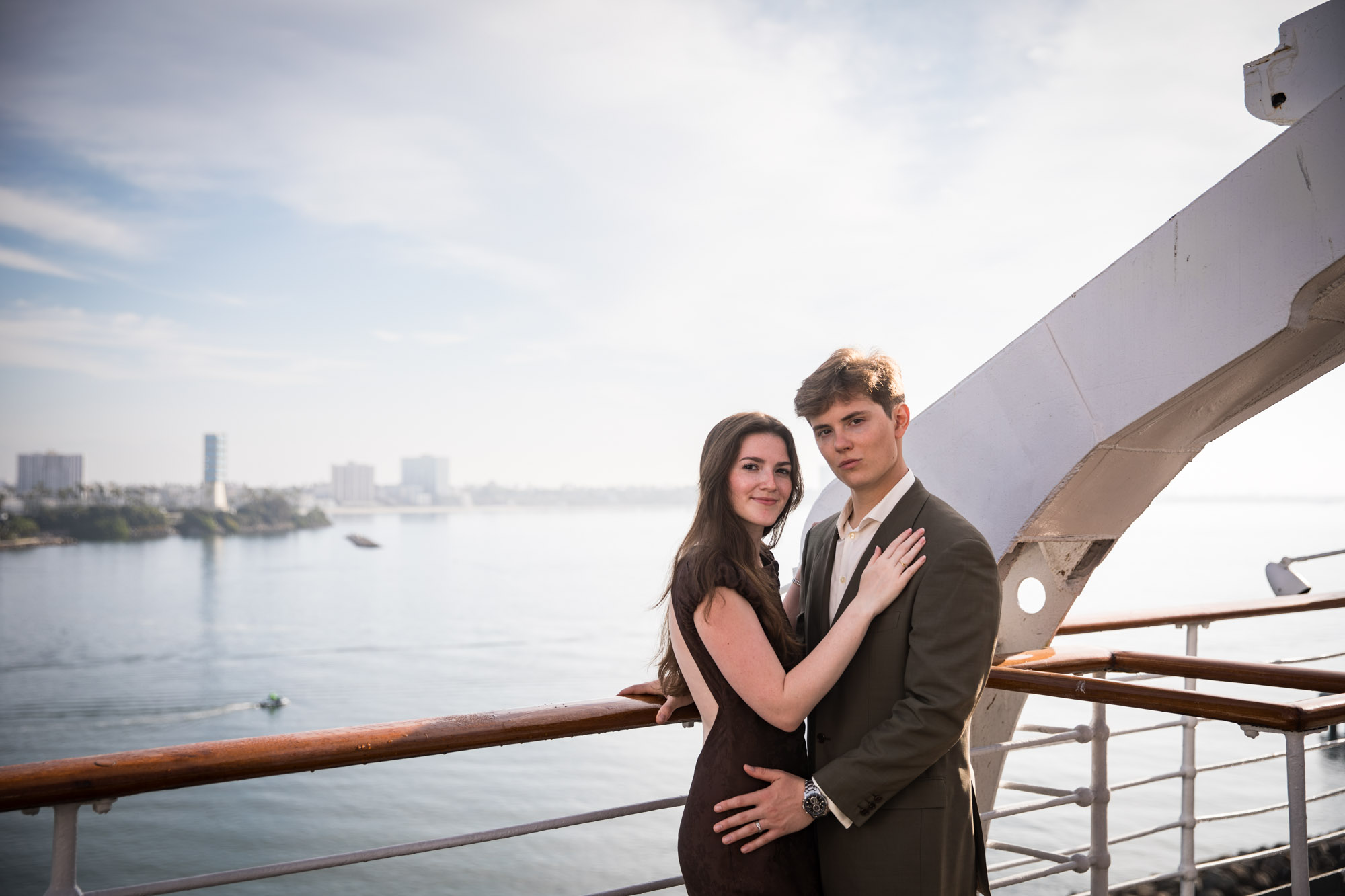 Couple embracing at the rail and looking at the camera, with a white gantry and the harbor behind them, photographed by a Long Beach photographer