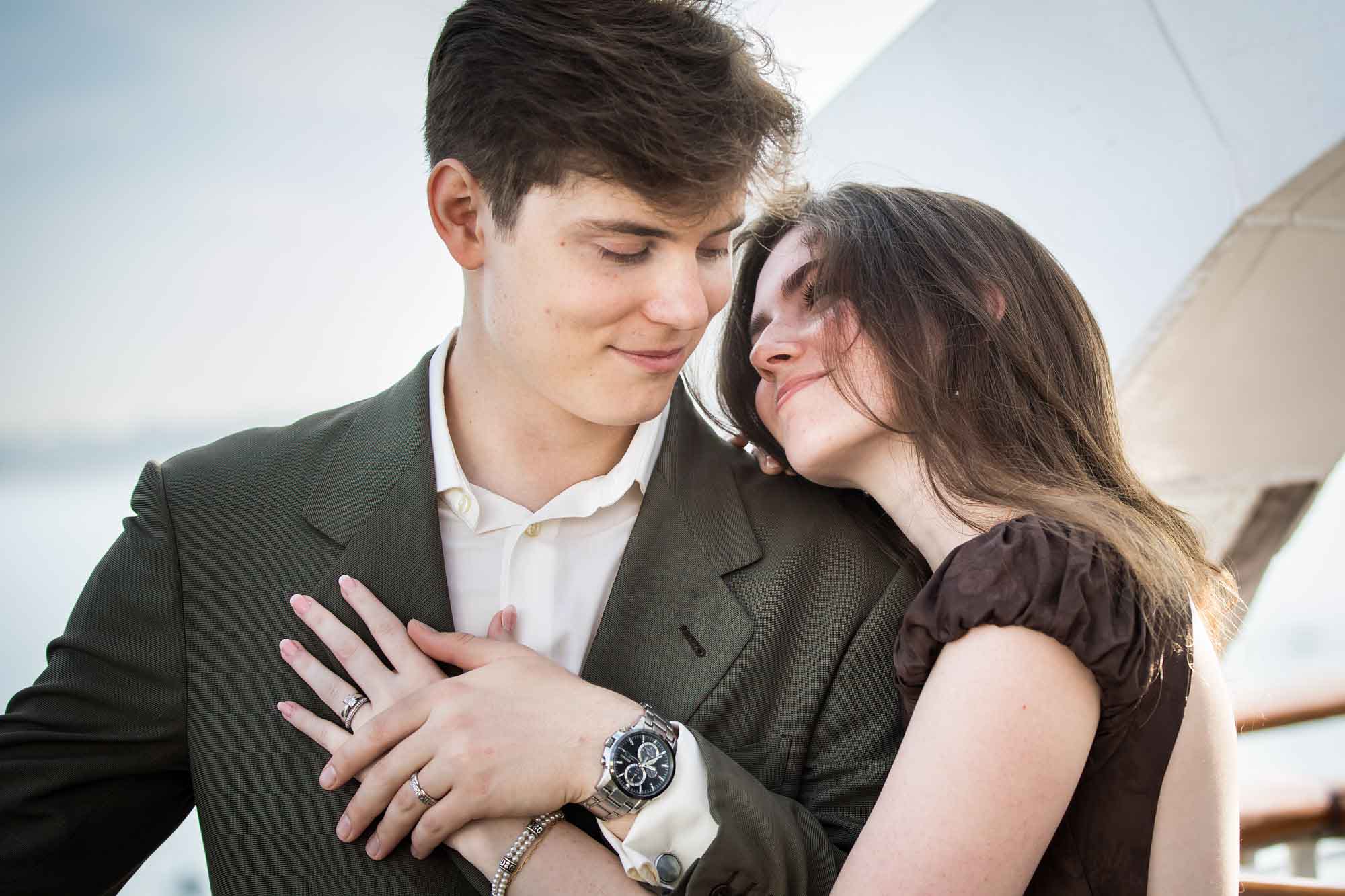 Close view of the woman resting against the man as he smiles down, her ring visible, on a Queen Mary photo shoot