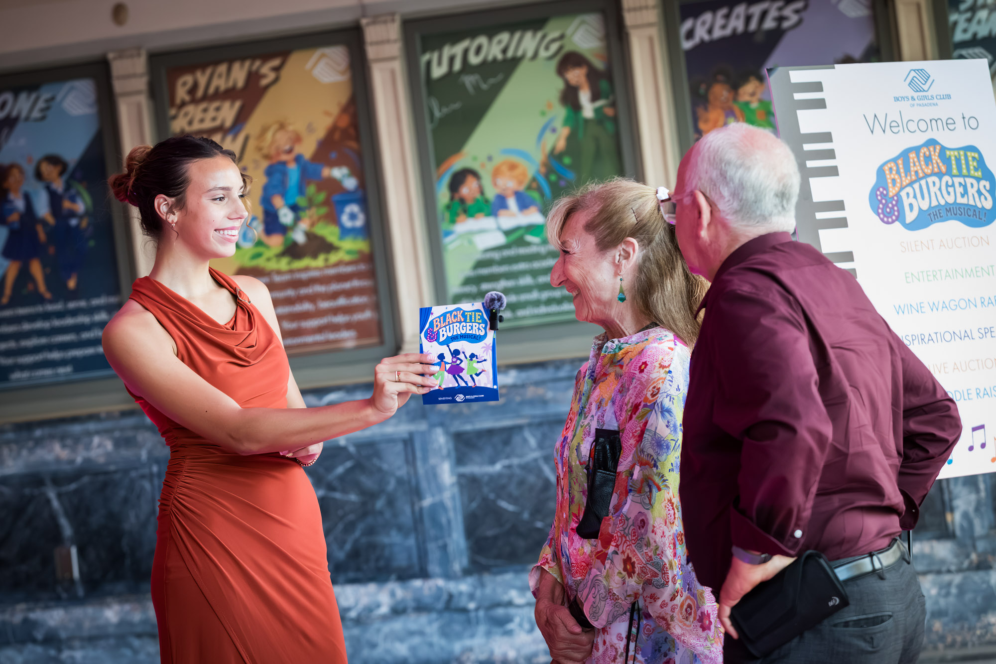 Raymond Theatre photos of a greeter showing the Black Tie & Burgers program to a smiling couple at the entrance