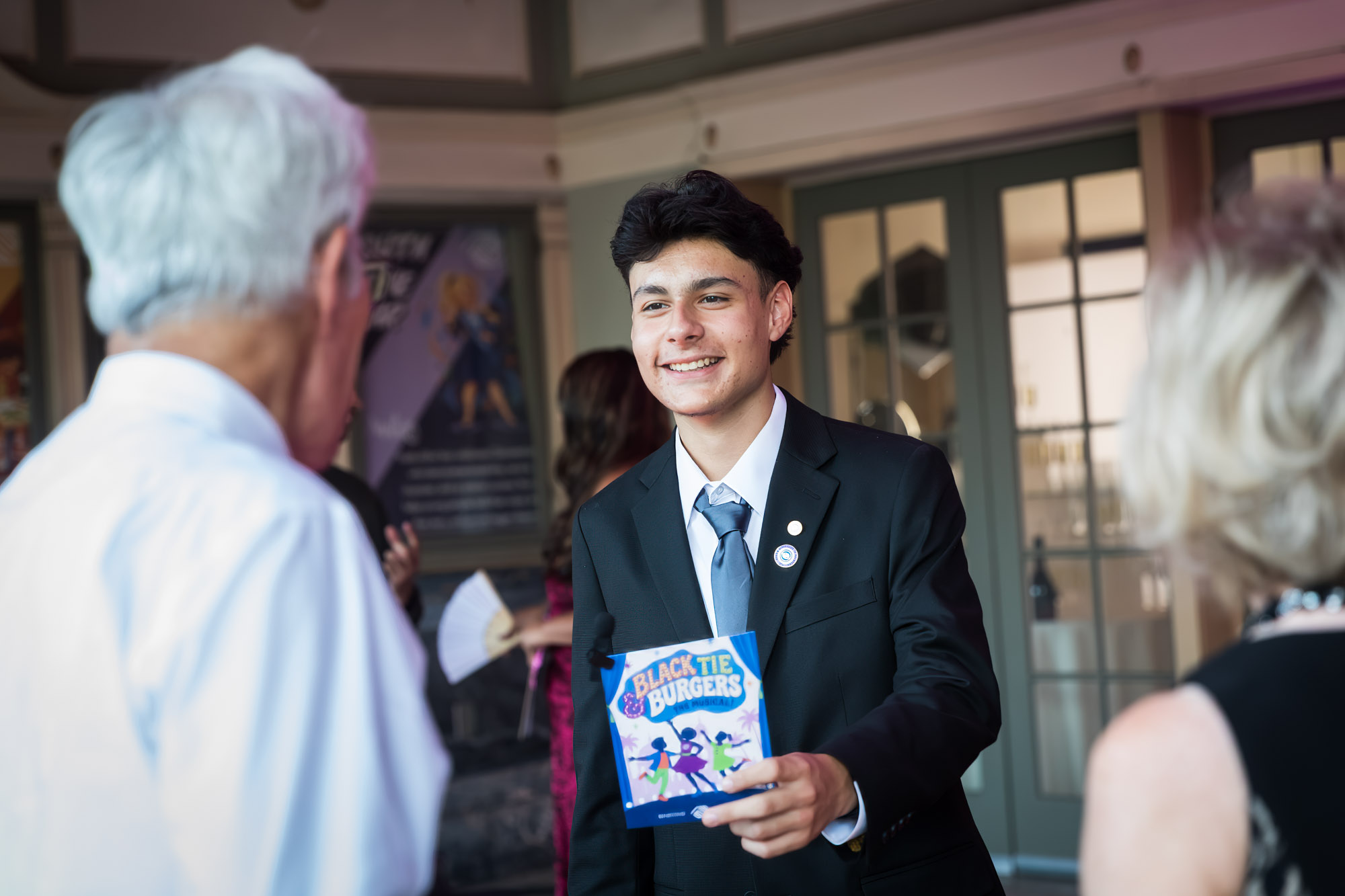 Raymond Theatre photos of a teen greeter in a suit handing out Black Tie & Burgers programs at the entrance
