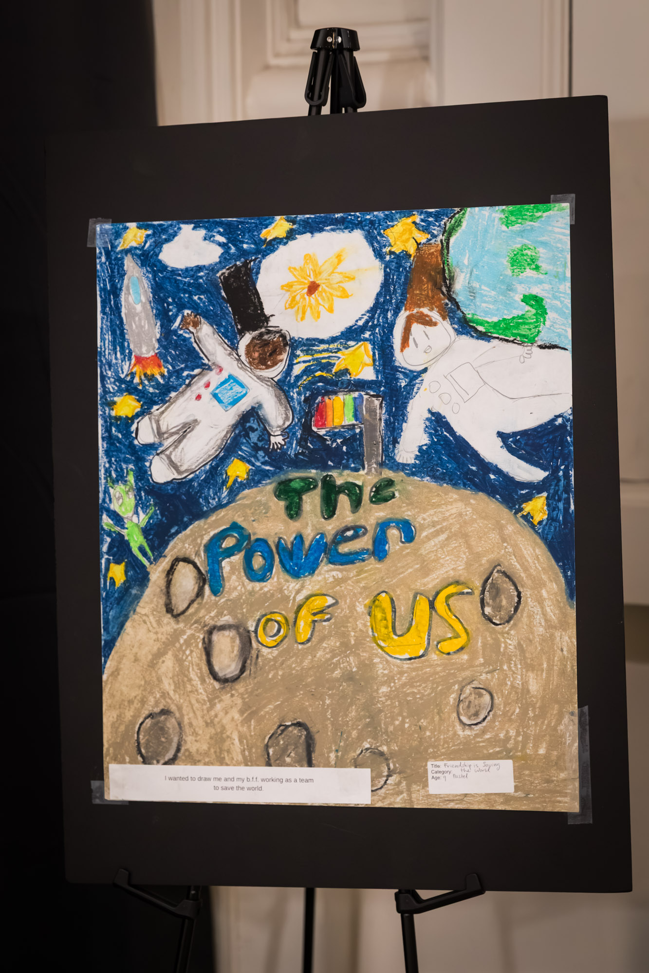 Raymond Theatre photos of a child's pastel artwork of two astronauts, titled The Power of Us, on an easel