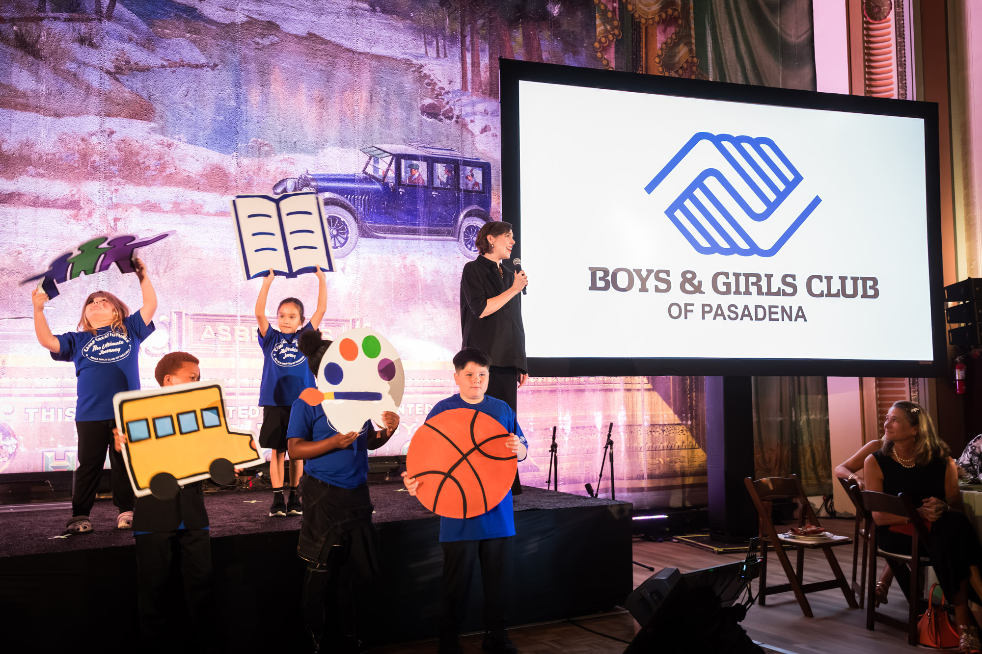 Raymond Theatre photos of kids in blue shirts holding giant book, bus, palette, and basketball cutouts on stage