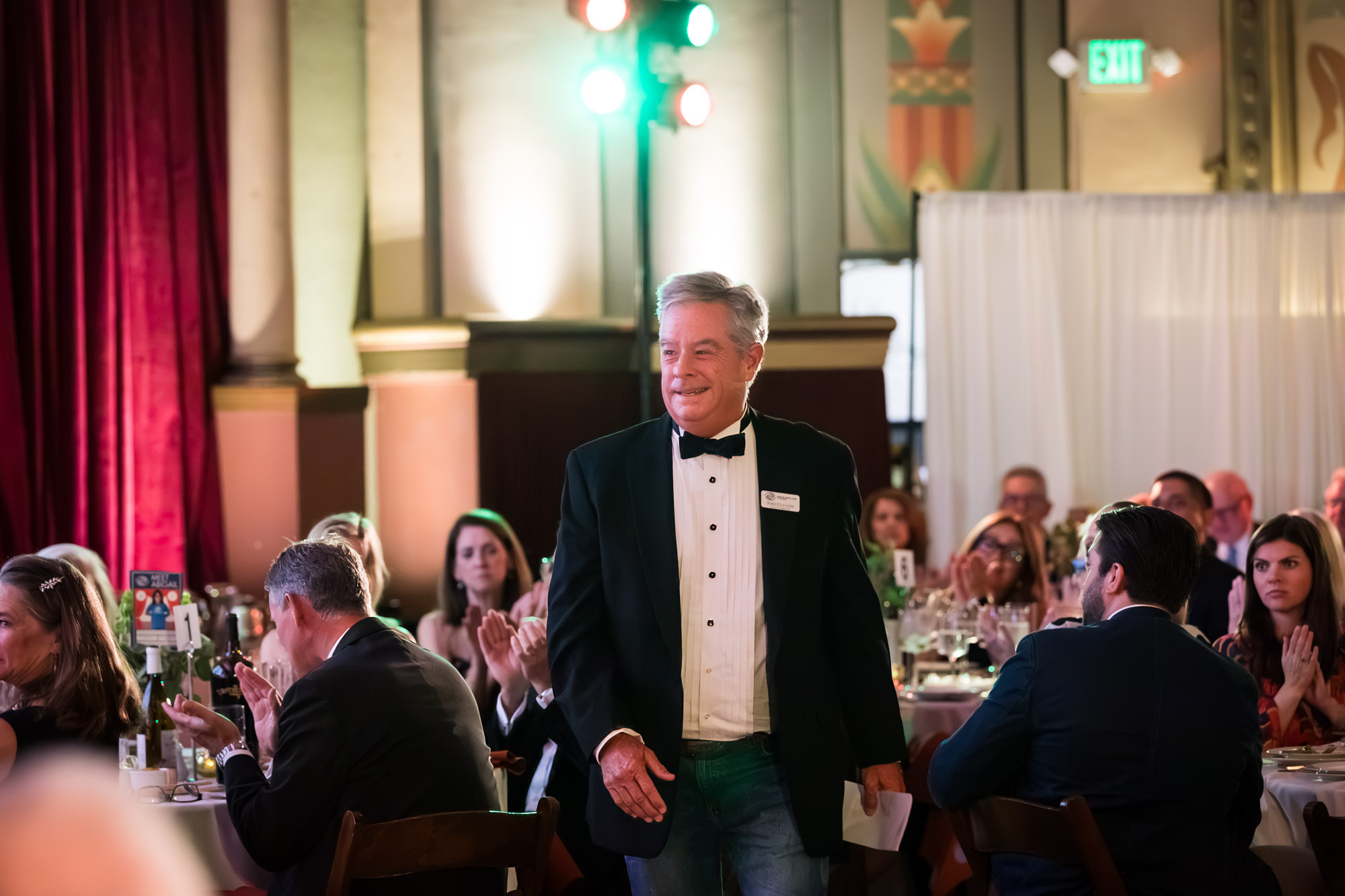 Raymond Theatre photos of a man in a tuxedo walking between tables as seated guests applaud