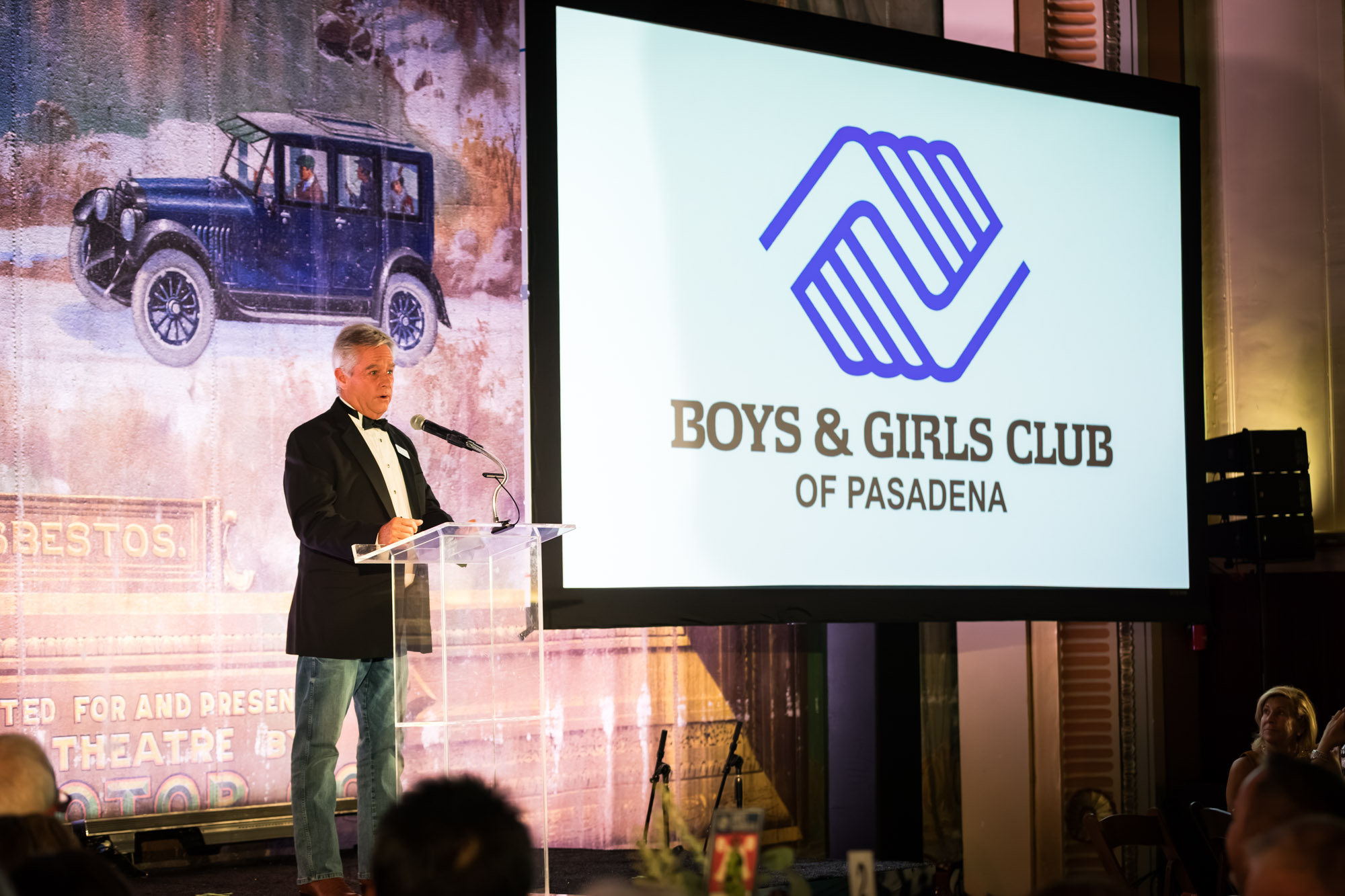 Raymond Theatre photos of a speaker in a tuxedo at a clear podium beside the Boys & Girls Club logo screen