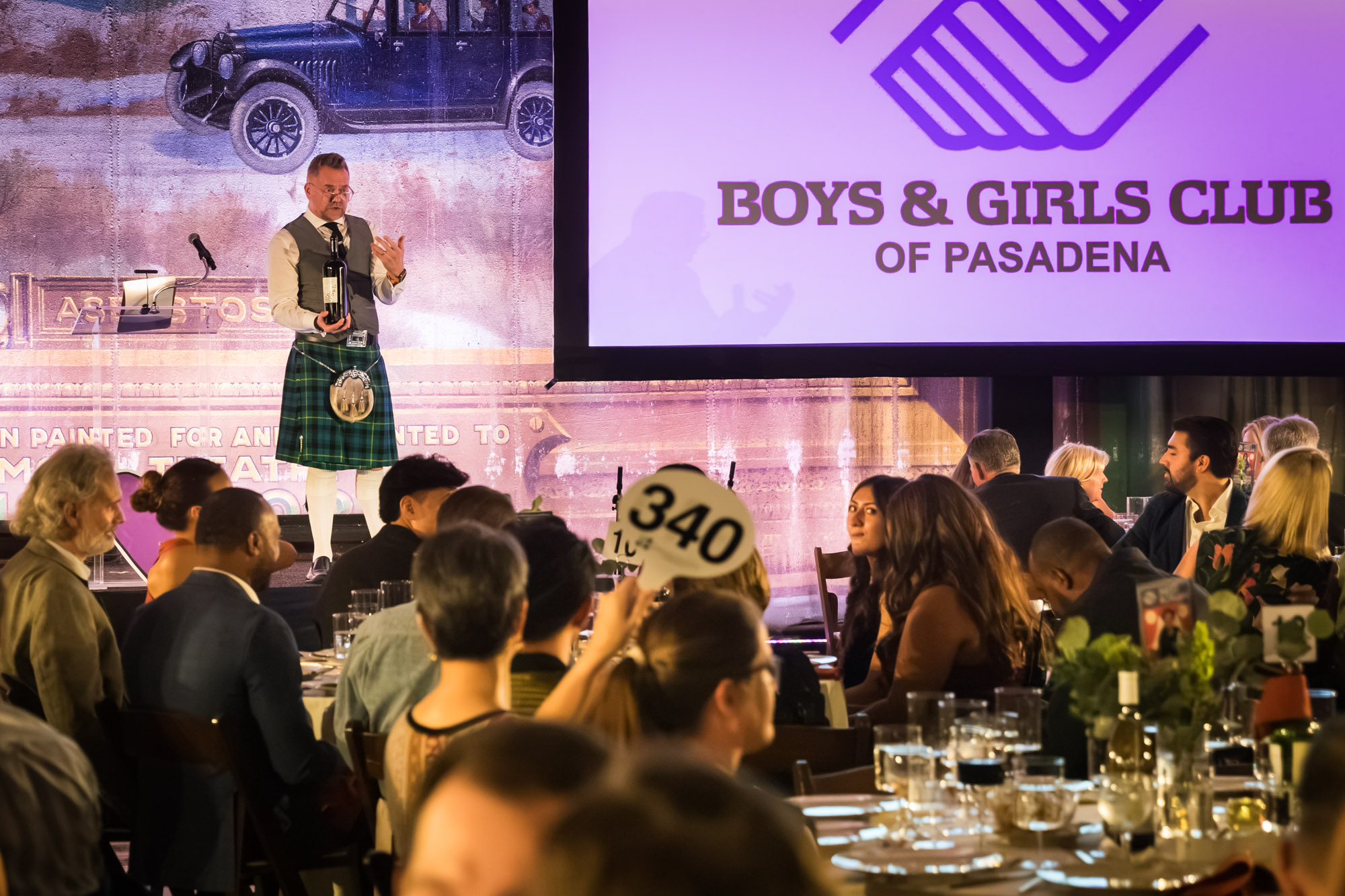 Raymond Theatre photos of a speaker in a kilt holding a wine bottle as a guest raises bidder paddle 340