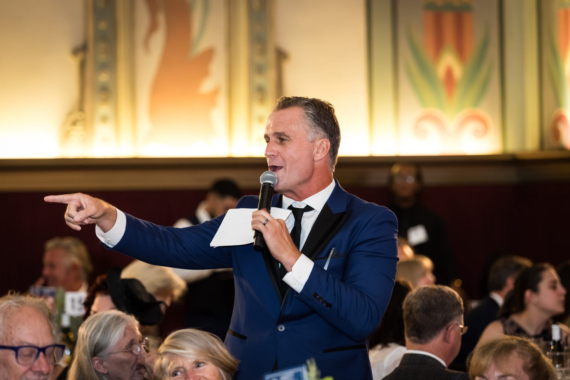 Raymond Theatre photos of a speaker in a blue tuxedo jacket pointing into the dining room with a microphone