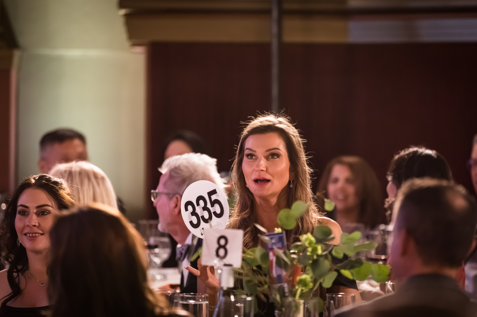 Los Angeles event photographer portrait of a woman holding a bidder paddle with a surprised smile at her table