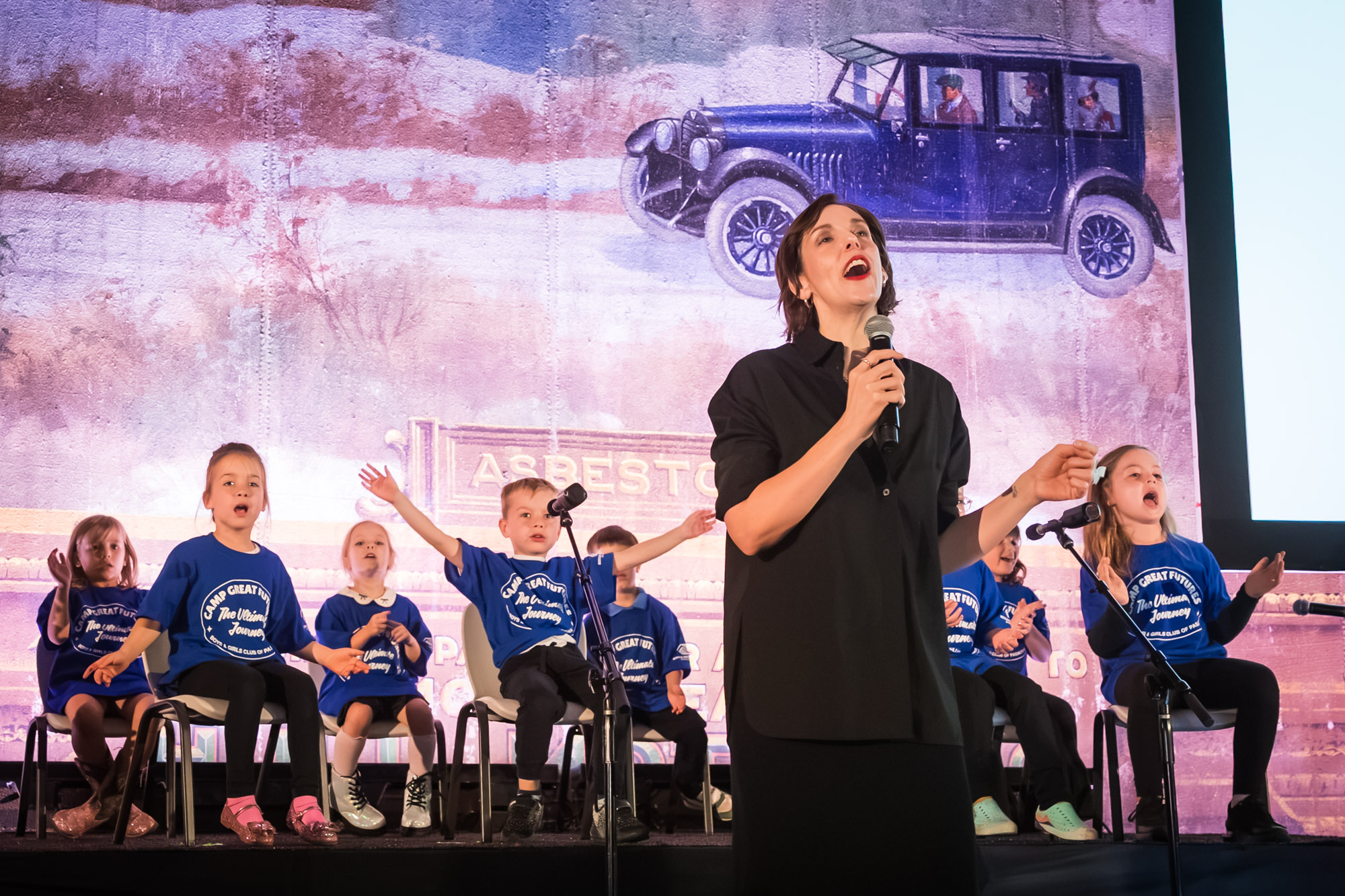 Raymond Theatre photos of a singer in black performing as kids in blue camp T-shirts clap along behind her