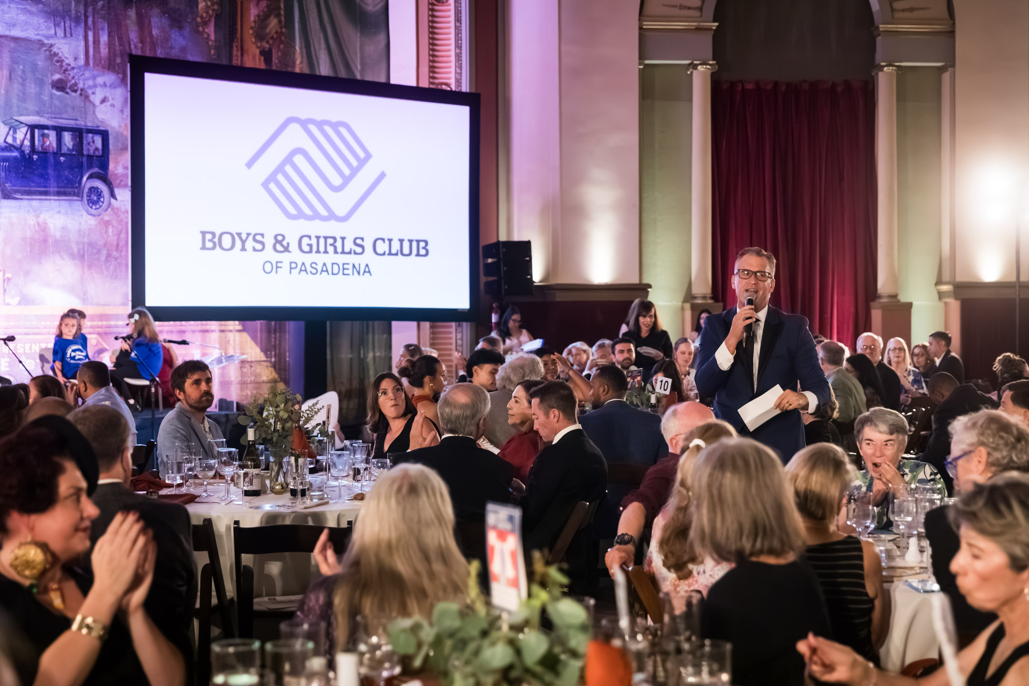 Los Angeles event photographer image of a speaker in glasses standing among dinner tables as guests applaud